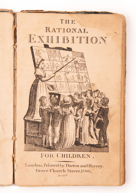 Ten young children gather around a large visual learning aid composed of forty pictures of everyday scenes arranged on a grid. A neatly dressed woman stands perched above the children on a chair. She holds a book in one hand and a pointer in the other, which she uses to direct the children's attention to one of the images. 